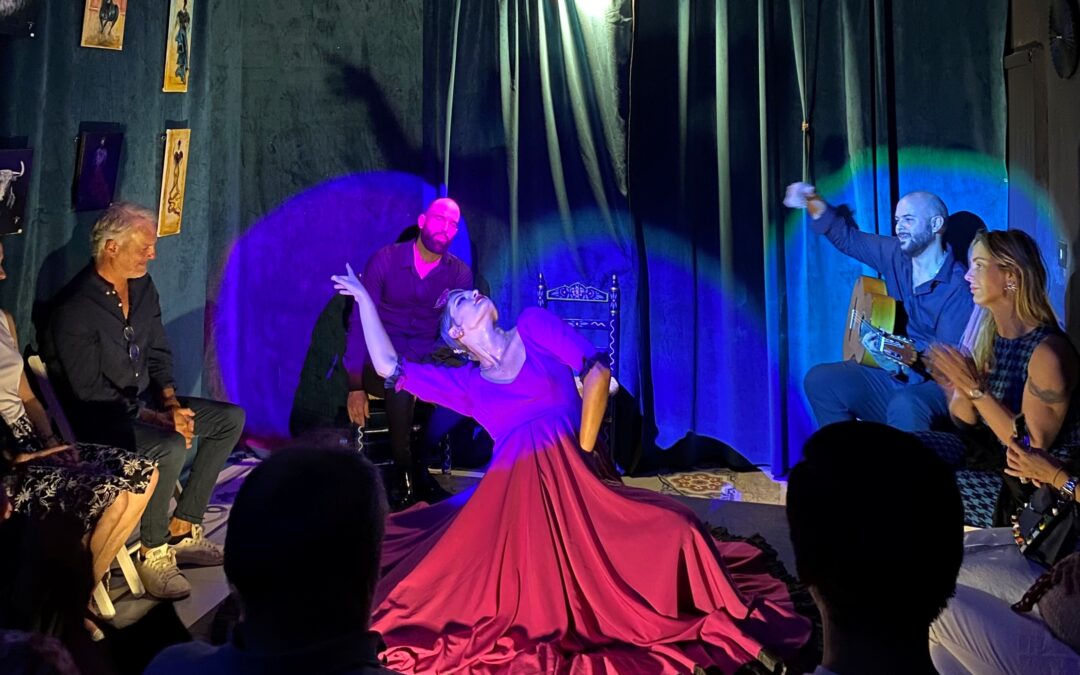 Bailarina en un escenario íntimo, vistiendo un vestido morado, realizando una pose elegante mientras el guitarrista toca en el fondo, con luces suaves iluminando la escena