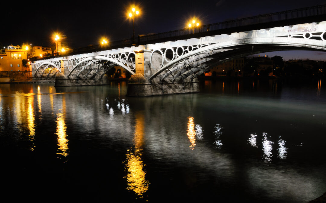 El Puente de Triana en Sevilla iluminado por la noche, reflejado sobre el río Guadalquivir.